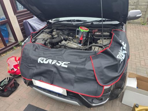 Car with open hood, covered with a black work mat, tools and equipment nearby.