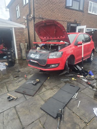 A red car with the bonnet open, surrounded by tools and parts on a driveway.