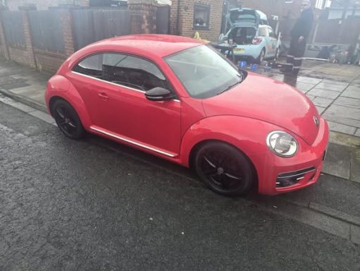 Red Volkswagen Beetle parked on a textured street with black alloy wheels.